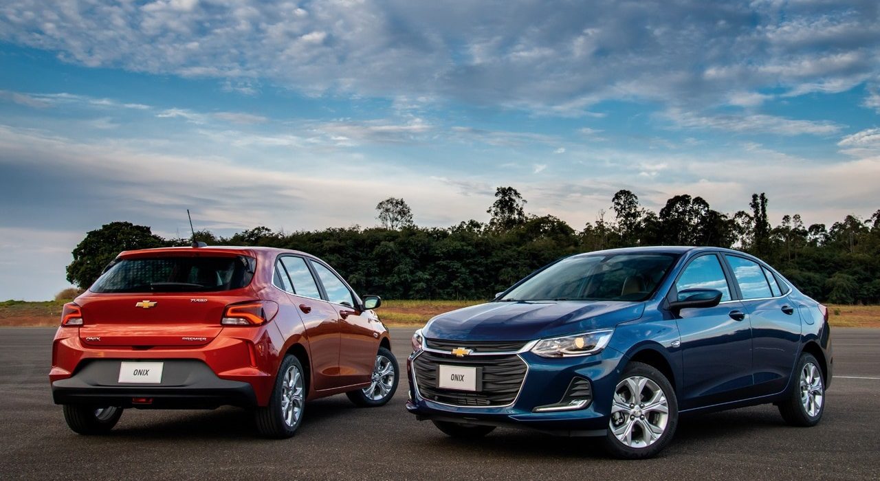 Dois modelos do Chevrolet Onix, um de frente e outro de ré. Um modelo é vermelho e o outro azul